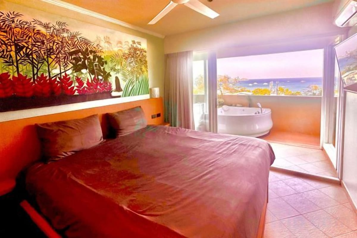 A bedroom with a large bed, a view of the sea, and a balcony with a hot tub.