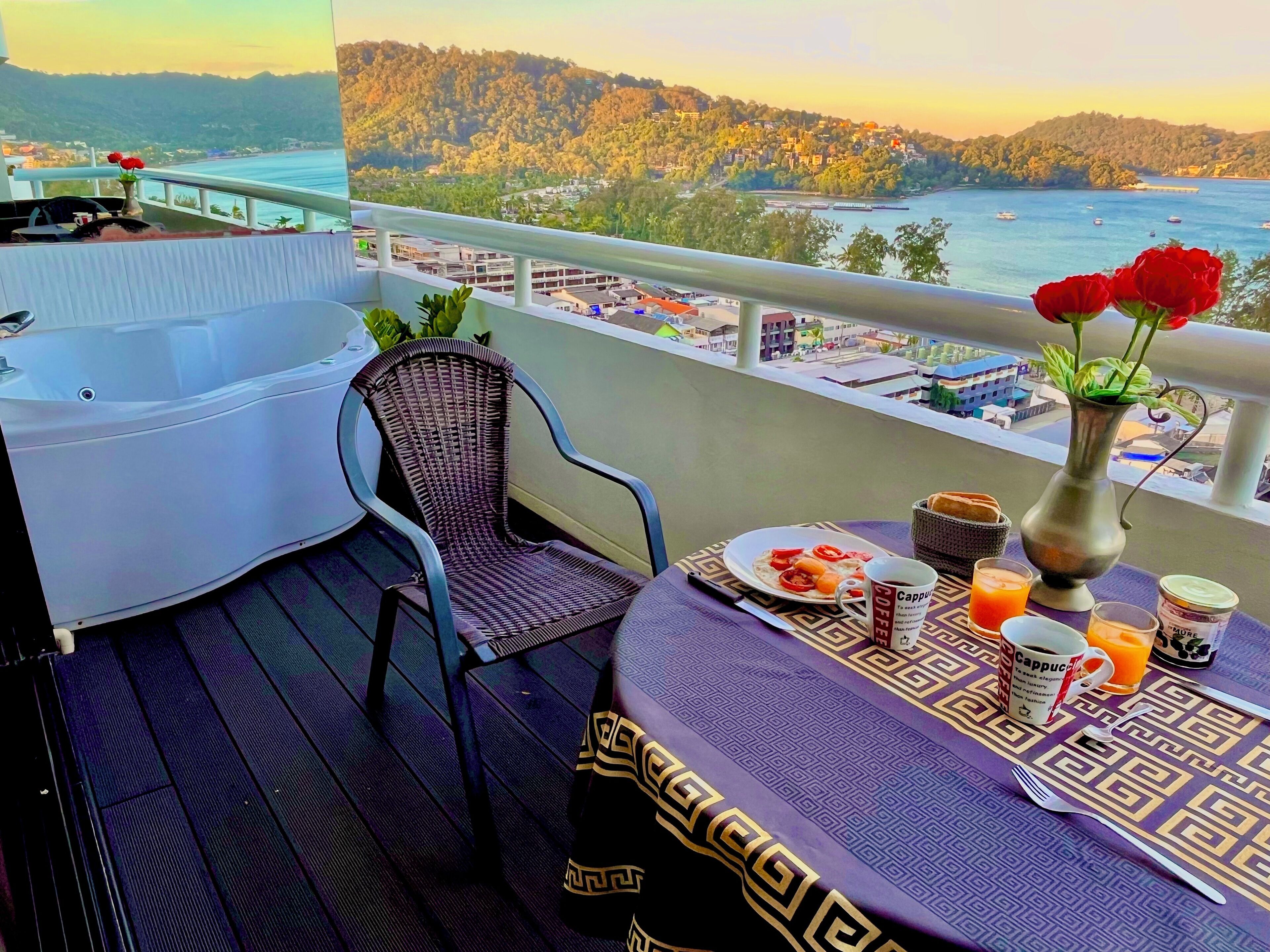 Uma varanda com vista para uma cidade costeira, uma cadeira de vime, uma mesa com itens de café da manhã e um vaso com flores.