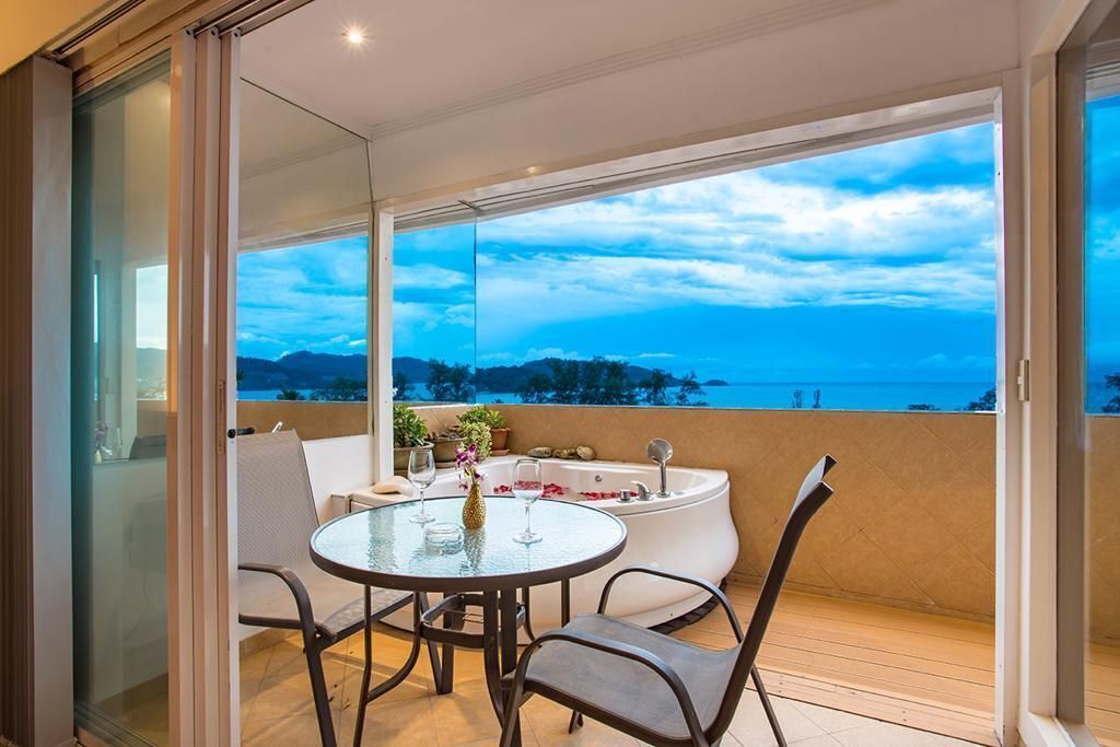 A balcony with a glass door, a round table with chairs, a bathtub, and a view of the ocean.