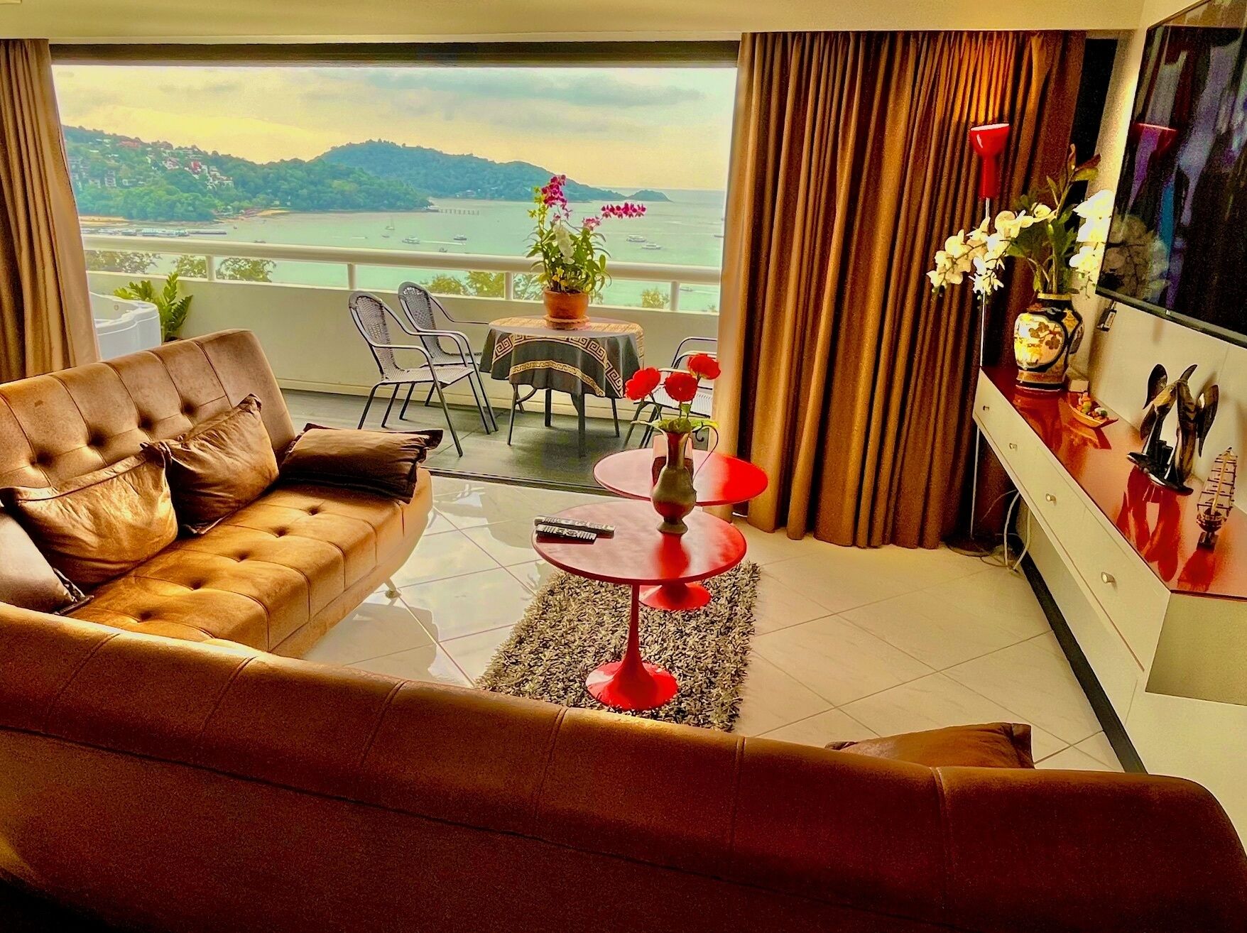 Sala de estar moderna com sofá de couro, uma mesa redonda pequena com um vaso de flores e uma TV em um móvel. Uma varanda com vista para o mar e colinas é visível pela janela.