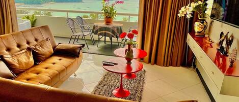 Sala de estar moderna com sofá de couro, uma mesa redonda pequena com um vaso de flores e uma TV em um móvel. Uma varanda com vista para o mar e colinas é visível pela janela.