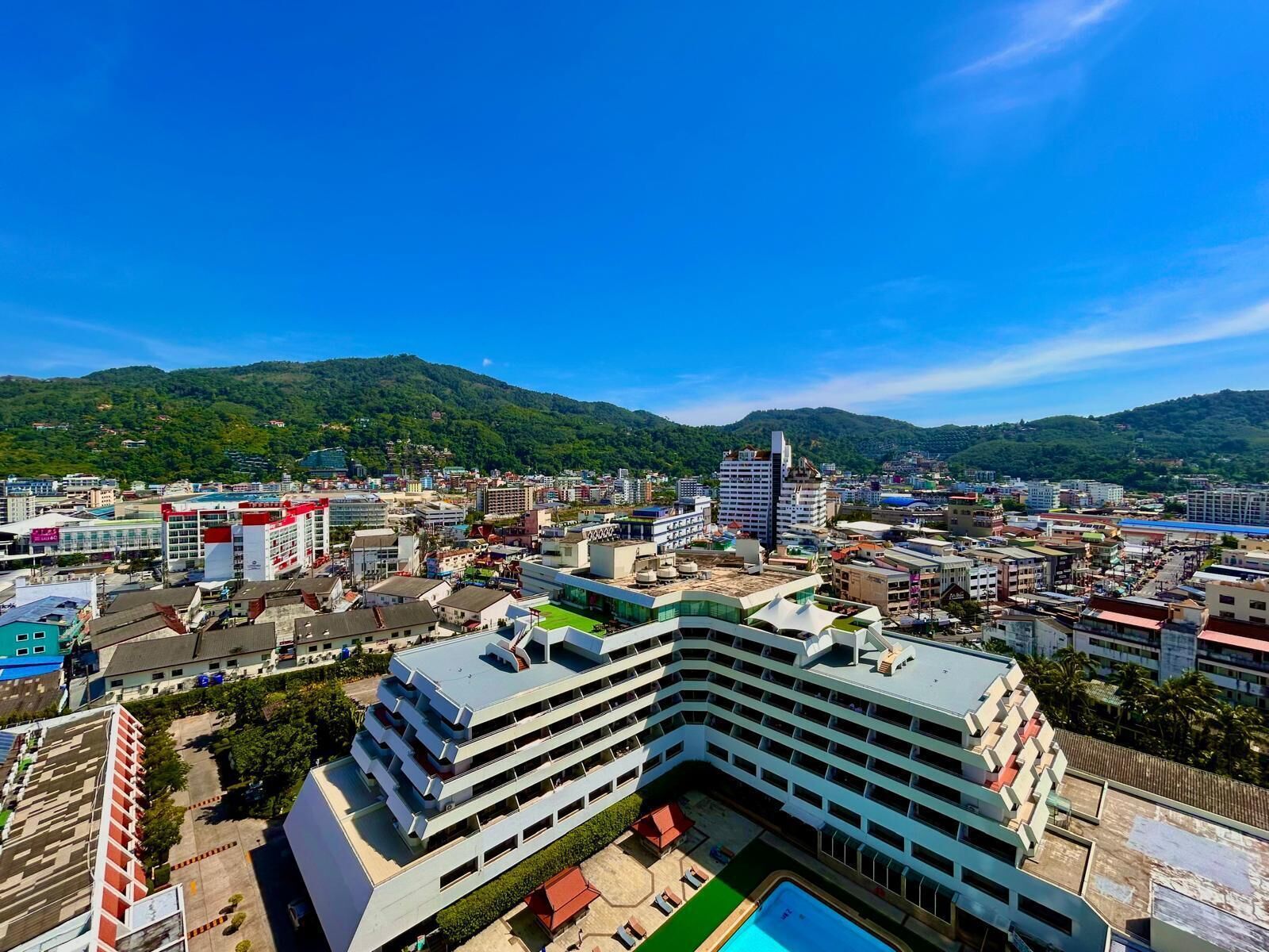 A multi-story hotel with a pool, surrounded by urban buildings and green hills in the background.