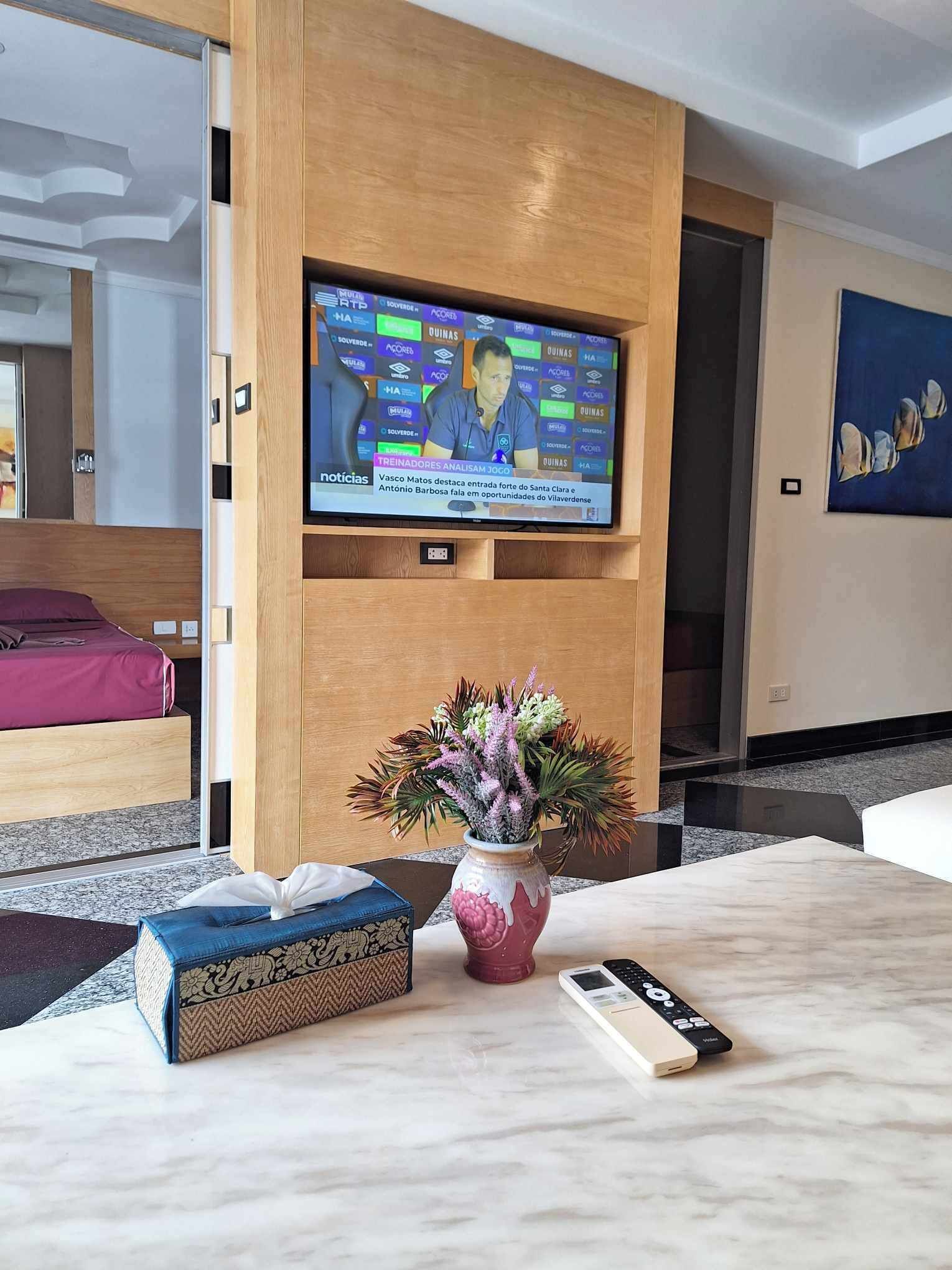 Ein Hotelzimmer mit einem an einer Holzplatte montierten Fernseher, einem Bett mit violettem Kopfteil, einer Blumenvase, einer Fernbedienung und einer dekorativen Kiste auf dem Boden.
