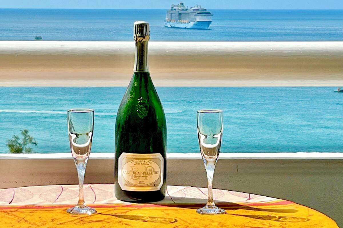 Ein Tisch mit einer Champagnerflasche und zwei Gläsern, mit Blick auf einen Strand und ein Kreuzfahrtschiff in der Ferne.