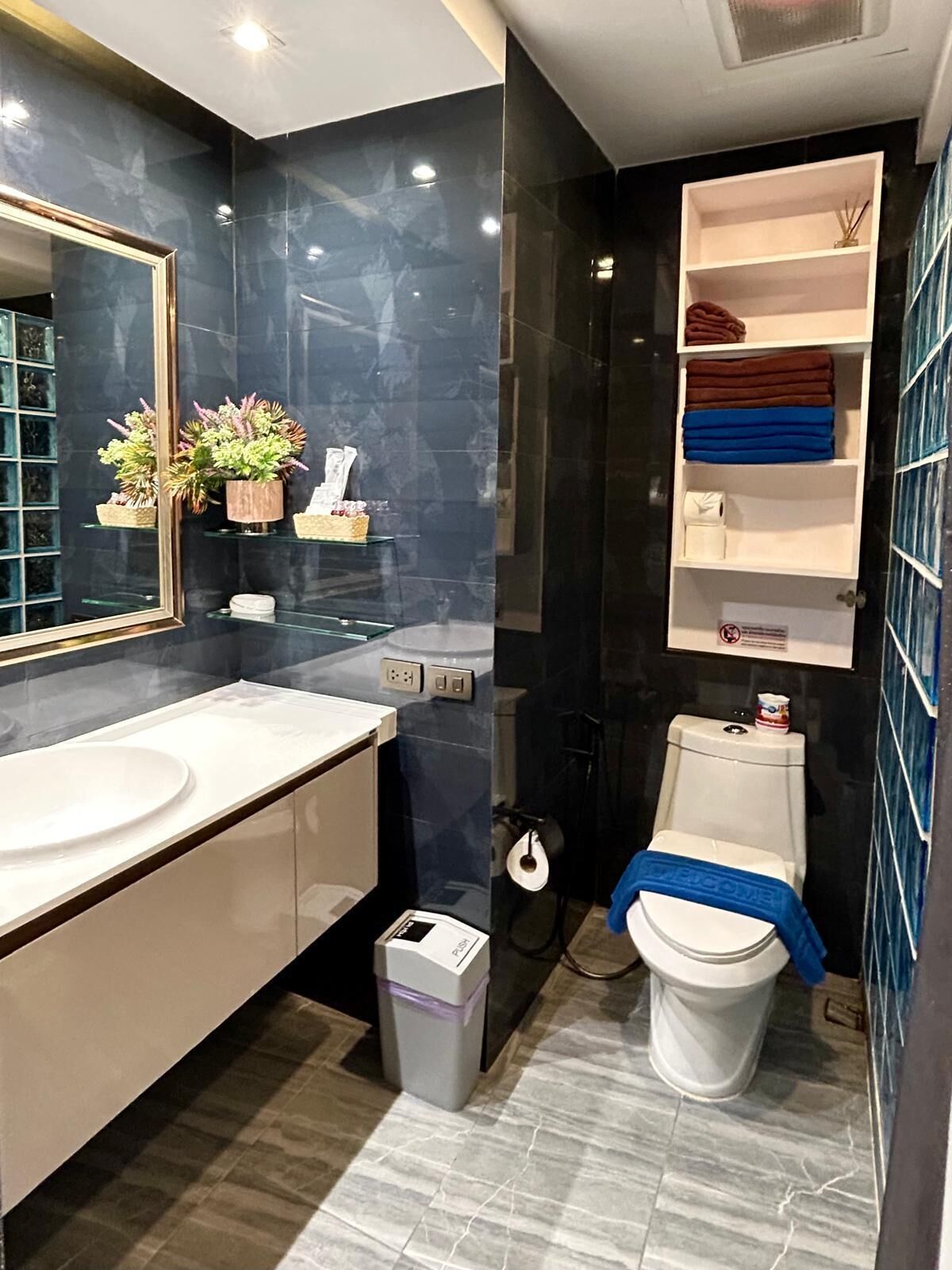 A modern bathroom with a large mirror, white sink, and a toilet with a blue towel. There are shelves with toiletries and a trash bin.