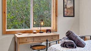 A bedroom with a wooden desk, a lamp, a clock, and a view of trees through a large window. - Schweitzer Lux Powder Dreams I Hot Tub I Families (Sandpoint)