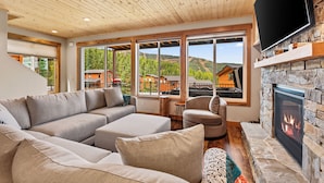 A modern living room with a large sectional sofa, a fireplace, and large windows offering a view of the mountains. - Schweitzer Lux Powder Dreams I Hot Tub I Families (Sandpoint)