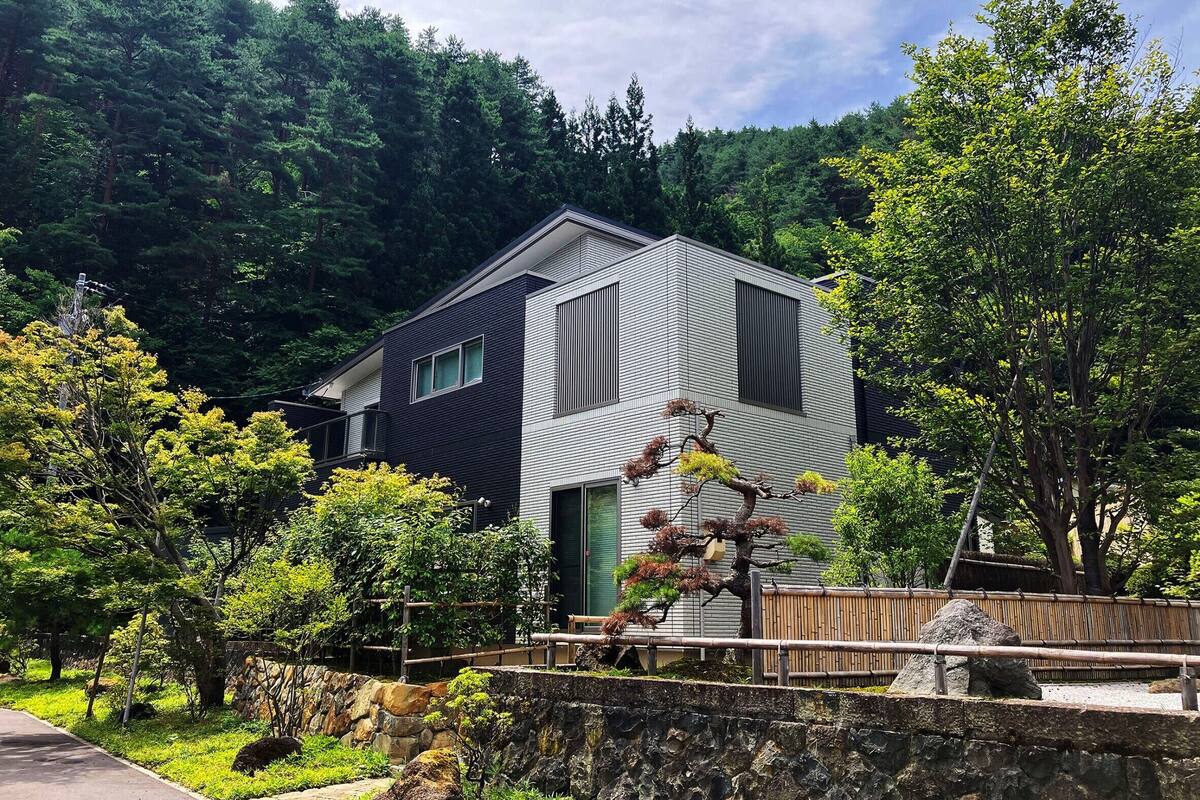 A modern two-story house with a dark exterior and large windows, surrounded by lush greenery and trees.