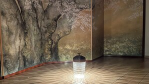 Une pièce avec un plancher en bois, un mur orné d’une fresque représentant un arbre et une petite table avec une lampe.