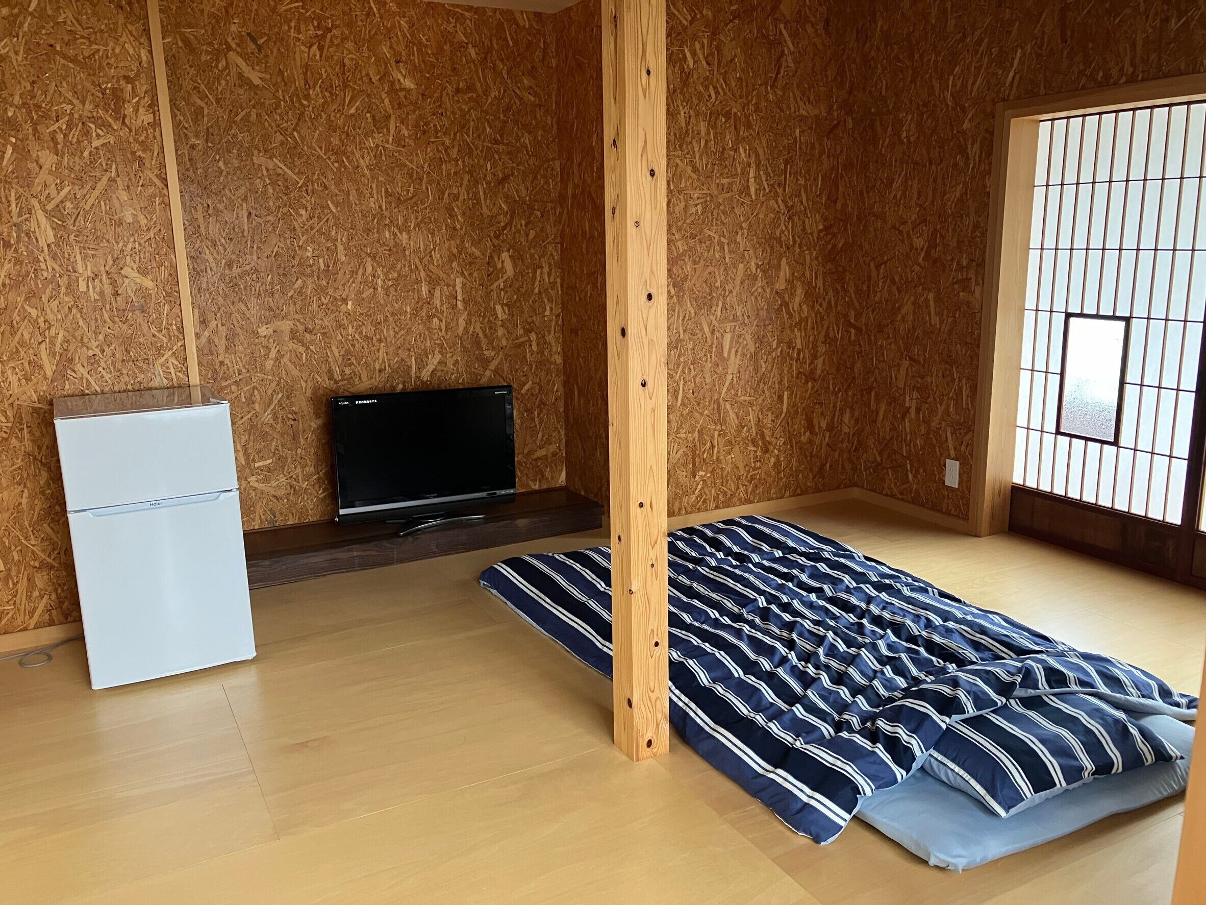 Una habitación con piso de madera, una cama con ropa de cama a rayas, un refrigerador blanco pequeño y un televisor sobre un mueble de madera.