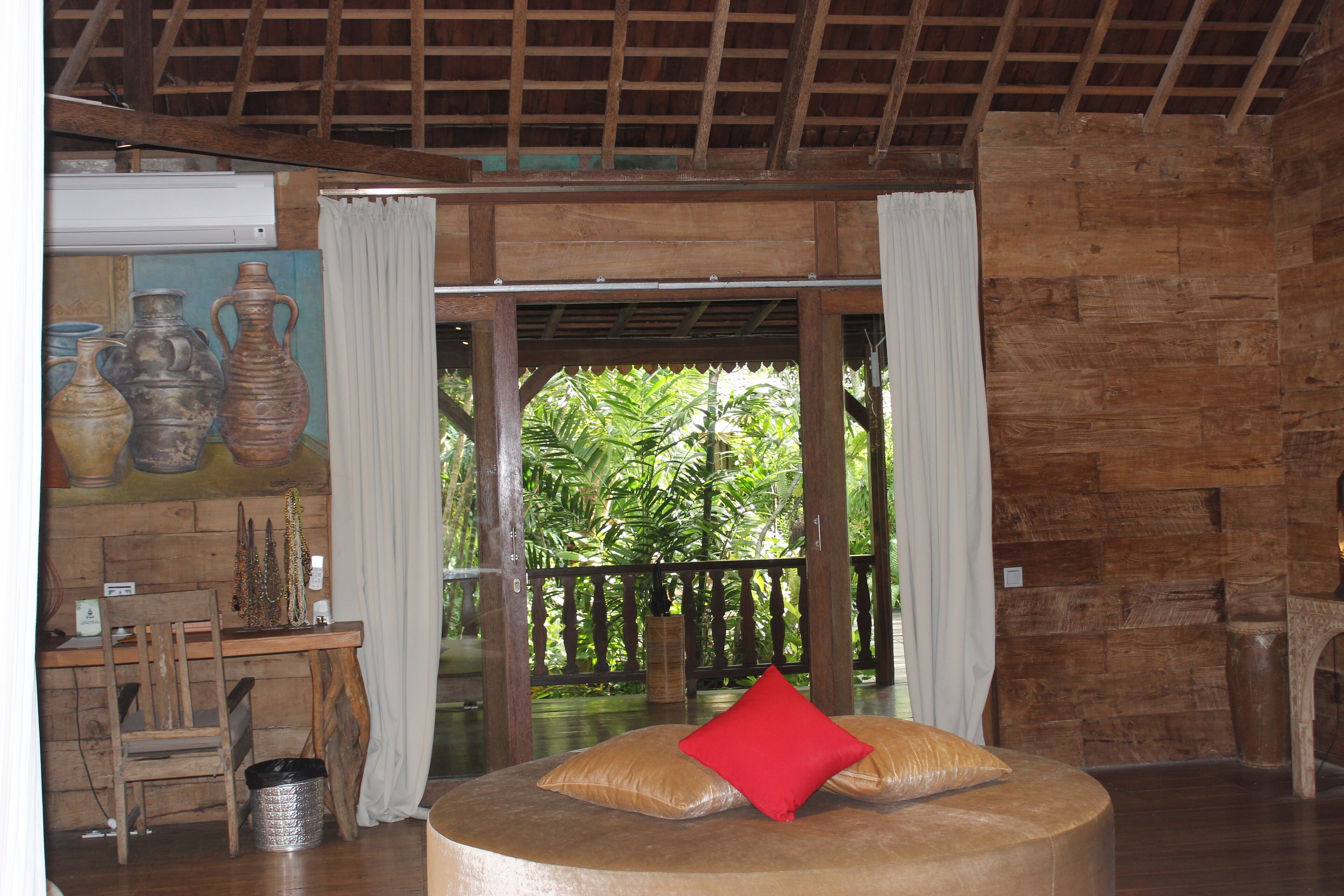 A room with a wooden ceiling, a round seating area with a red pillow, a wooden desk with a chair, and a view of lush greenery through a glass door.