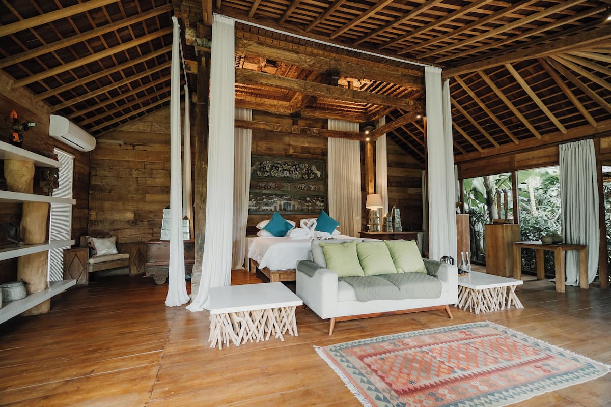 A spacious room with a wooden ceiling, a bed with pillows, a white sofa, a small table, and a patterned rug.