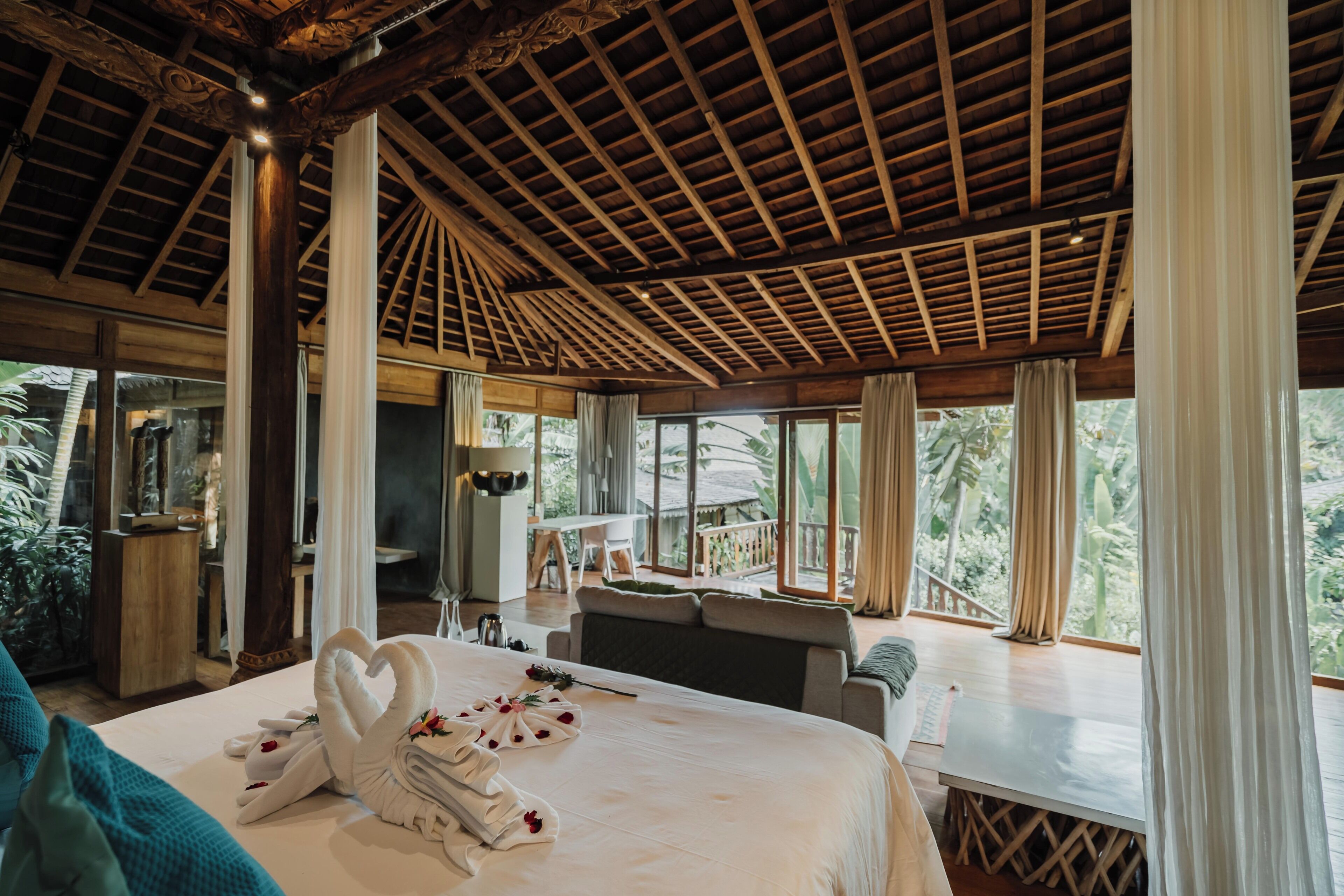 A spacious bedroom with a large bed, a balcony with a view of the outdoors, and a wooden ceiling structure.