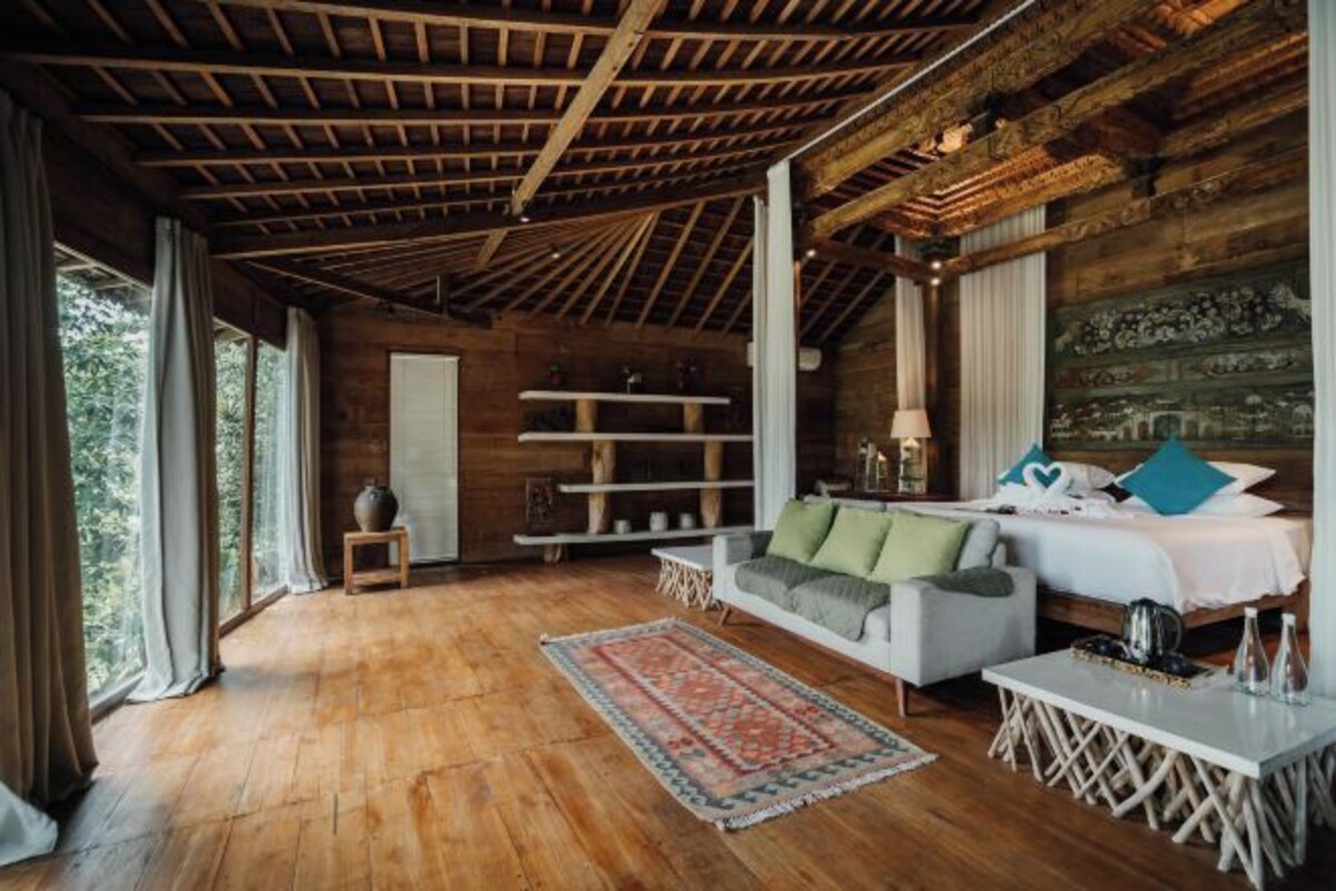 A spacious room with wooden flooring, a white sofa, a bed with green and blue pillows, and a large window with curtains.