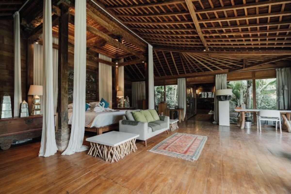 A spacious room with wooden flooring, a high ceiling with exposed beams, a sofa set, a dining table, and a view of greenery outside.