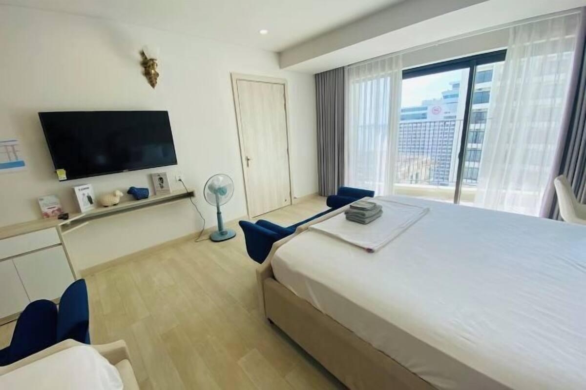 A hotel room with a bed, a flat-screen TV, a fan, a desk, and a window with curtains.