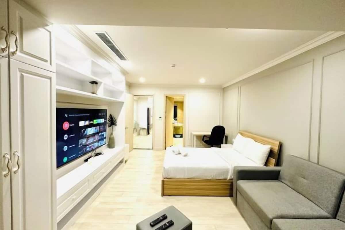 A modern hotel room with a flat-screen TV, a bed with a wooden headboard, a grey sofa, and a desk.