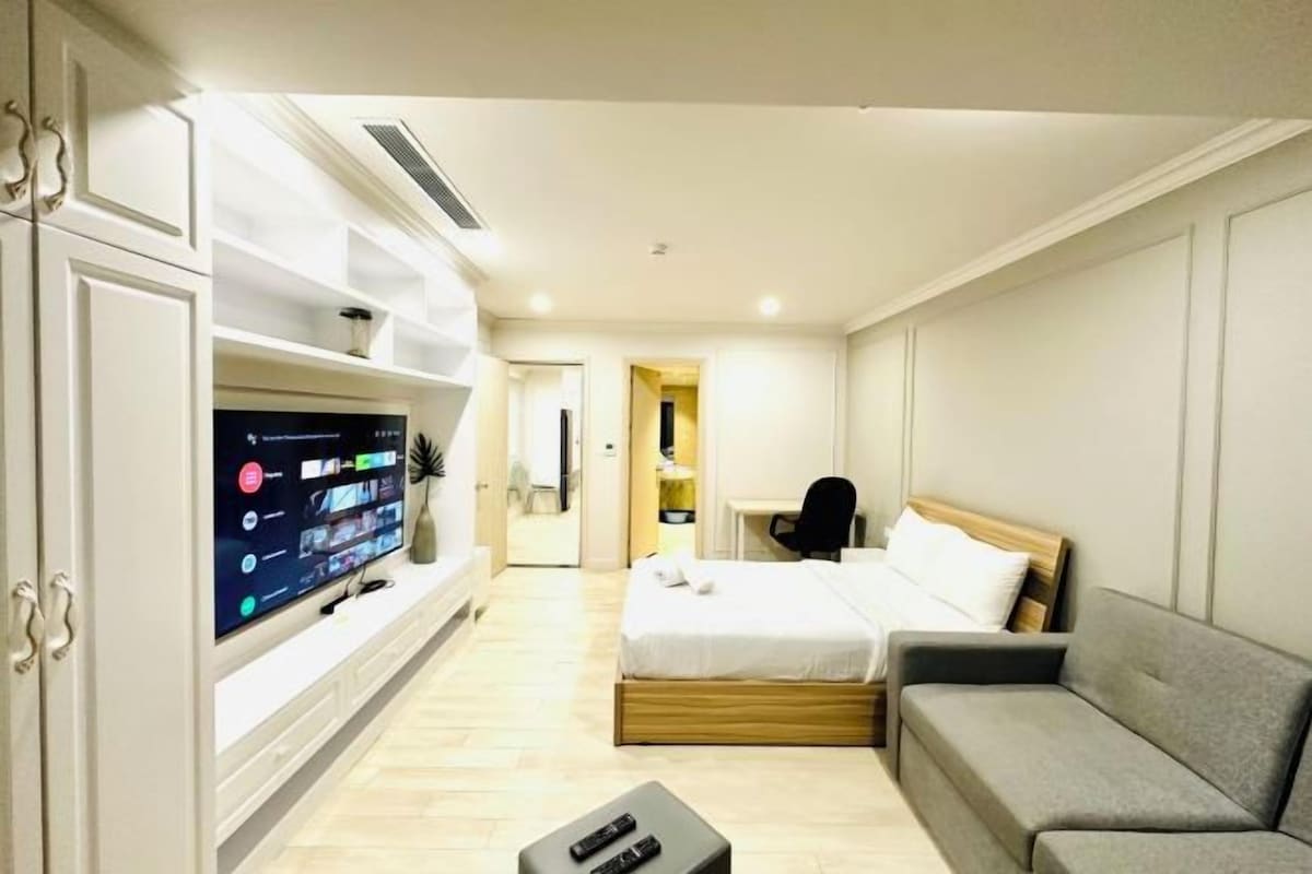 Ein modernes Hotelzimmer mit Flachbildfernseher, einem Bett mit Holz Kopfteil, einem grauen Sofa und einem Schreibtisch.