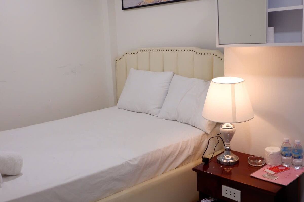 A neatly made bed with a white headboard, a bedside lamp, and a small wooden nightstand with a mirror and a few items on it.