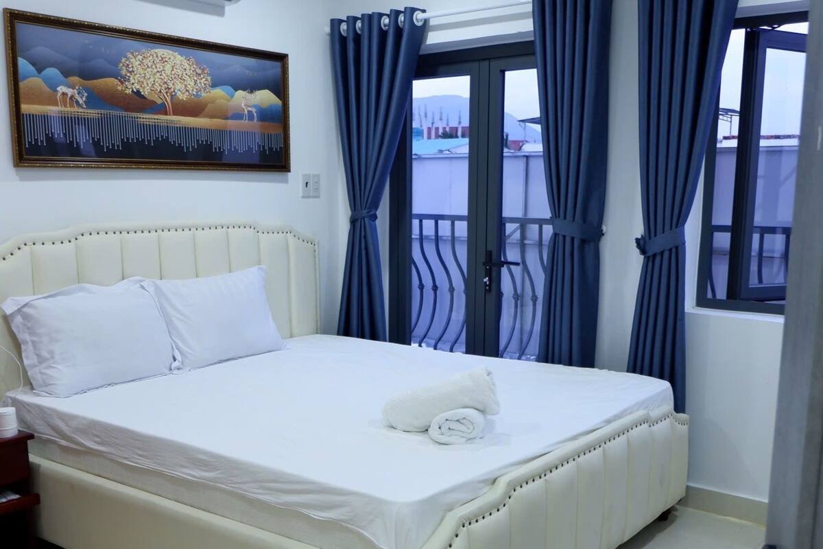 A hotel room with a bed, a painting on the wall, and a window with curtains.