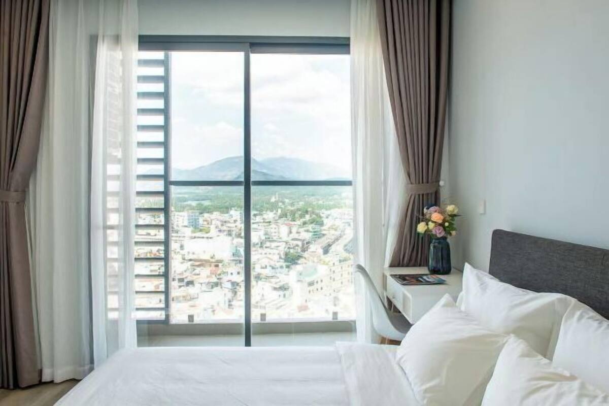 A bedroom with a large window, a bed, a bedside table with a vase of flowers, and a view of a cityscape.