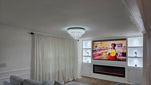 Ein modernes Wohnzimmer mit flachbildfernseher an der Wand, grauen Sofas, einer Kristallleuchter und einem weißen Vorhang.