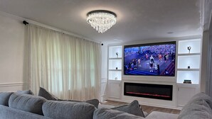 Ein Wohnzimmer mit einem großen Flachbildfernseher an einer weißen Wand, grauen Sofas, einer Kristallleuchtre und einem Fenster mit durchsichtigen Vorhängen.
