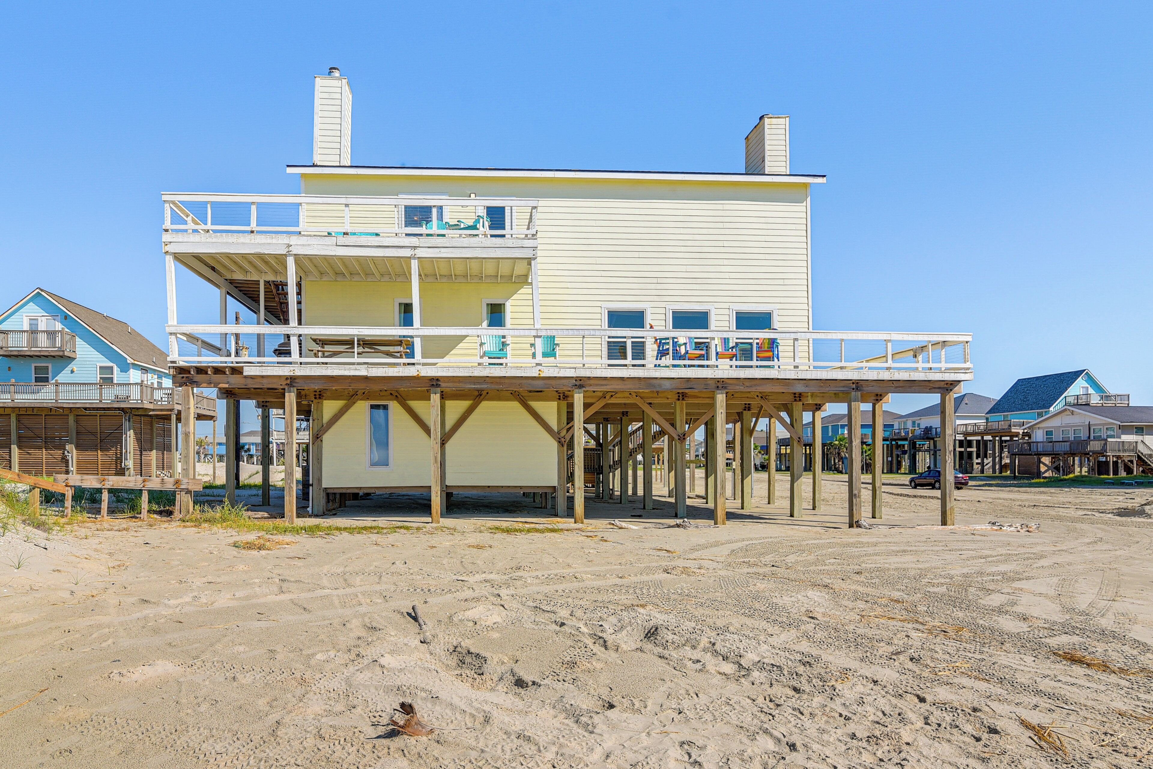 A two-story beachfront house with a balcony, a covered patio, and a sandy beach in the foreground.