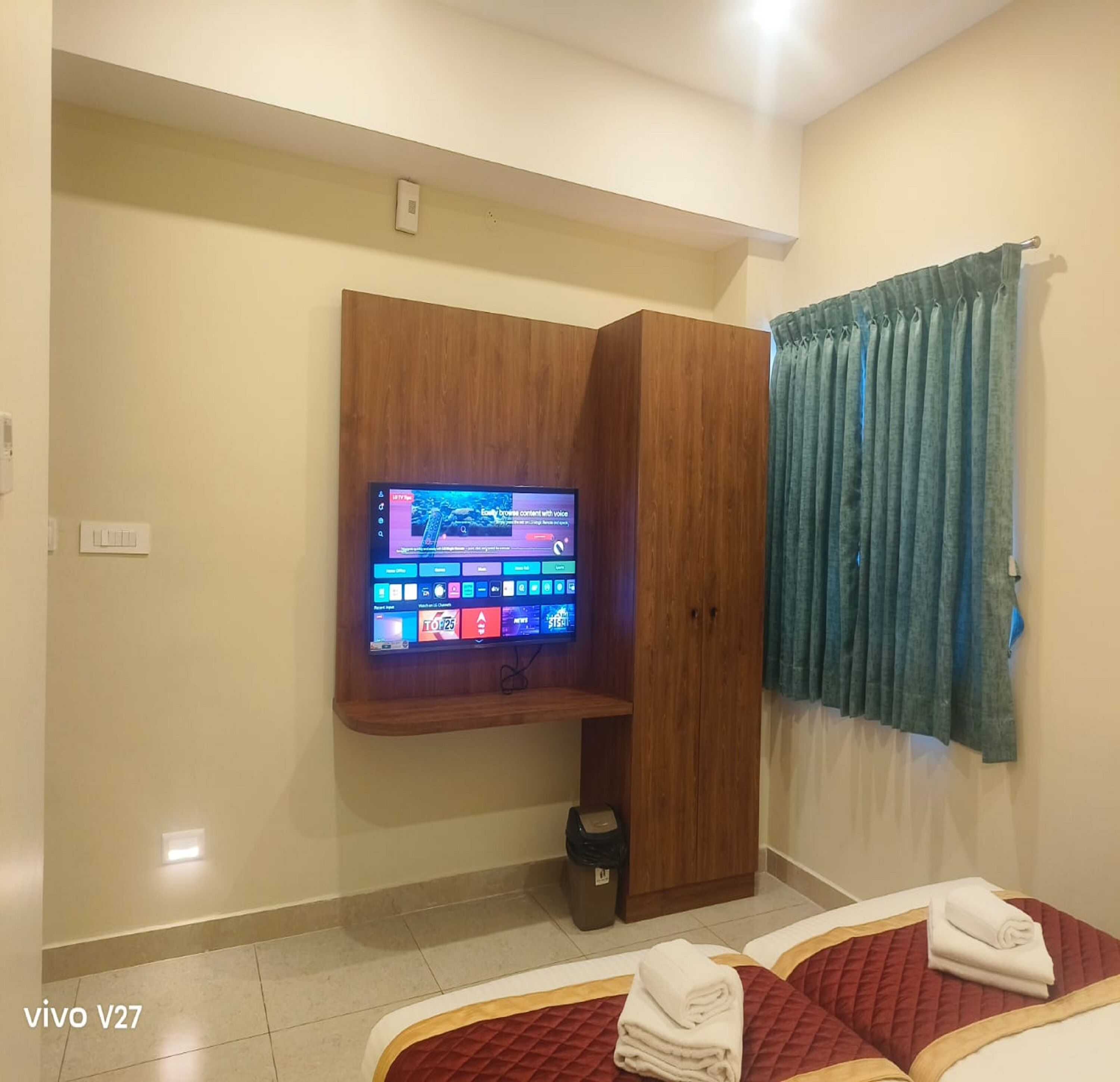 A hotel room with a TV mounted on the wall, a wooden wardrobe, a bed with a red quilt, and a blue curtain.