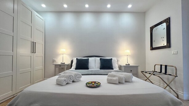 A neatly made bed with white linens and a folded towel, two bedside lamps, a small table with a lamp, a wall-mounted mirror, and a metal drying rack. - Aster apartment - Free Parking in the historic center of Alba - Langhe (Alba)