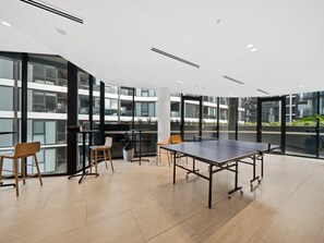 A modern indoor space with a ping pong table, wooden chairs, and large windows offering views of a building.