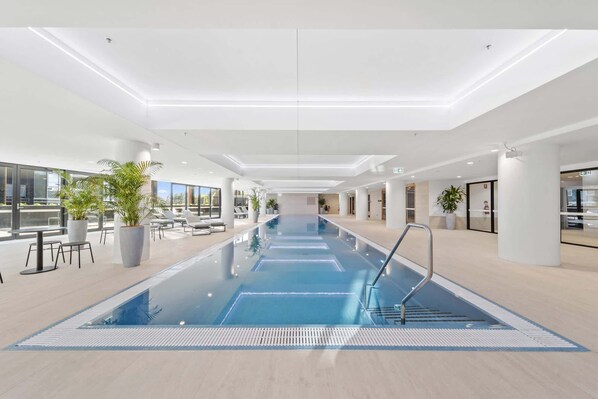 A modern indoor swimming pool with clear water, surrounded by lounge chairs and potted plants.