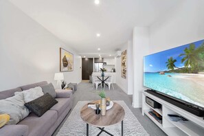 A modern living room with a flat-screen TV, a grey sofa, a coffee table, and a dining area with a white table and chairs.