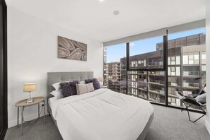 A modern hotel room with a large bed, a nightstand, a lamp, a chair, and a large window with a city view.