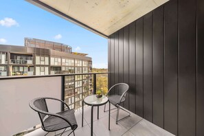 En balkon med to stole og et lille bord, udsigt til en bygning overfor, og en klar himmel.