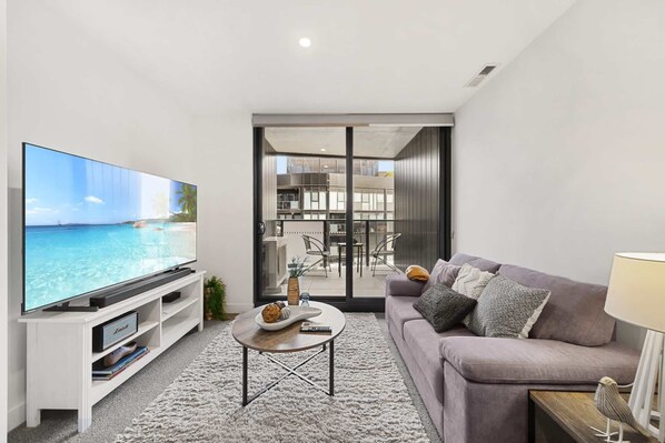 A modern living room with a flat-screen TV, a grey sofa, a coffee table, and a view of a balcony with outdoor furniture.