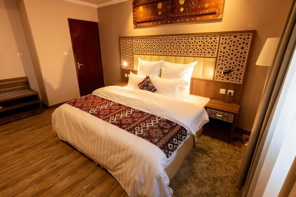 A hotel room with a large bed, a wooden bench, a decorative headboard, and a nightstand with a lamp.
