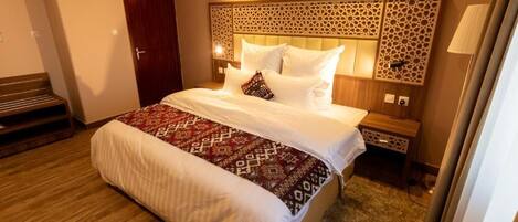 A hotel room with a large bed, a wooden bench, a decorative headboard, and a nightstand with a lamp.
