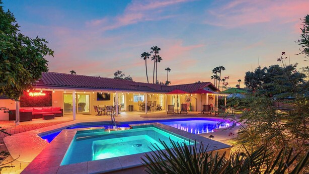 A pool area with a house in the background, surrounded by palm trees and outdoor seating. - The Rusty Cactus | Heated Salt Pool, Spa, & More! (Phoenix)