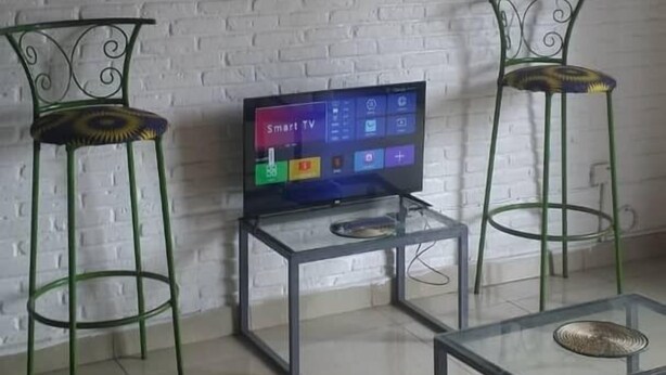 A living room with a white brick wall, a mounted TV, two green metal chairs, and a glass coffee table. - GreenPage Apartments Kiyovu  (Kigali)