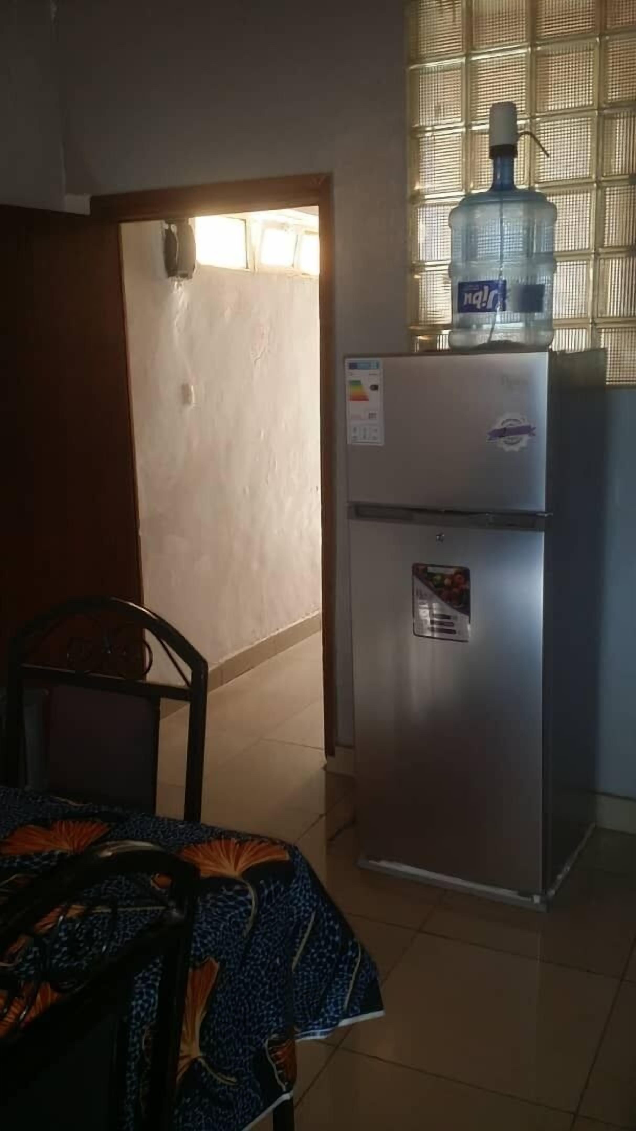 A kitchen with a refrigerator, a water dispenser, a dining table with a chair, and a window with blinds.