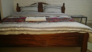 A wooden bed with a patterned bedspread and pillows, a small bedside table, and a white brick wall in the background. - GreenPage Apartments Kiyovu  (Kigali)