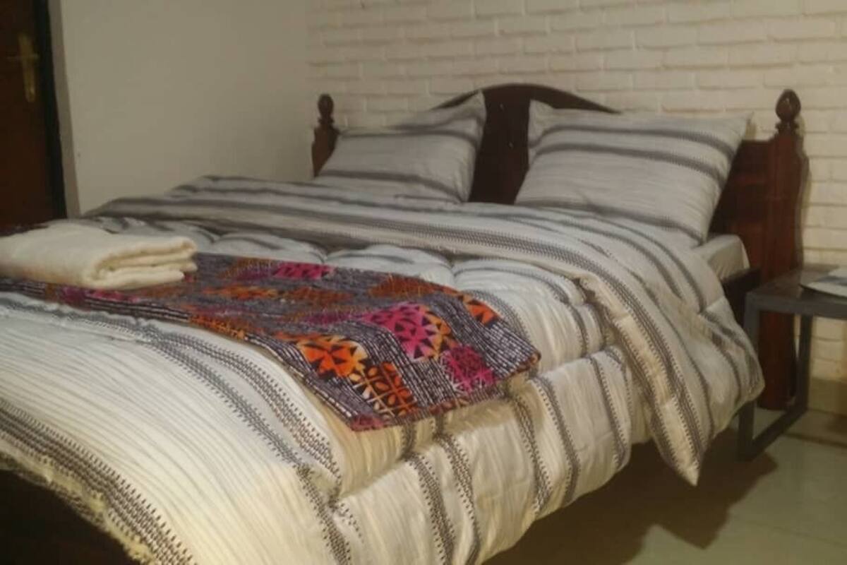 Un lit bien fait, avec une couverture rayée et un plaid coloré, une tête de lit en bois et une table de chevet avec une lampe.