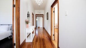 Un pasillo con suelo de madera, paredes blancas y un candelabro. Se ve una cama con ropa de cama blanca a la izquierda y una consola blanca con un jarrón de flores en el centro.