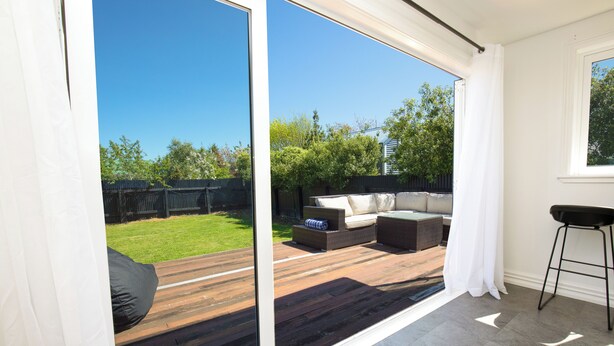 Una sala de estar moderna con un amplio ventanal corredera que da a una terraza de madera y un jardín.