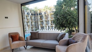Un salon avec un canapé et des fauteuils de couleur beige, une grande fenêtre donnant sur un immeuble d’hôtel et des palmiers.