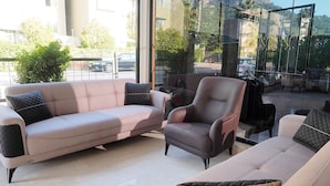 Un salon moderne avec un canapé, un fauteuil et un pouf. De grandes fenêtres offrent une vue sur la rue et les immeubles.