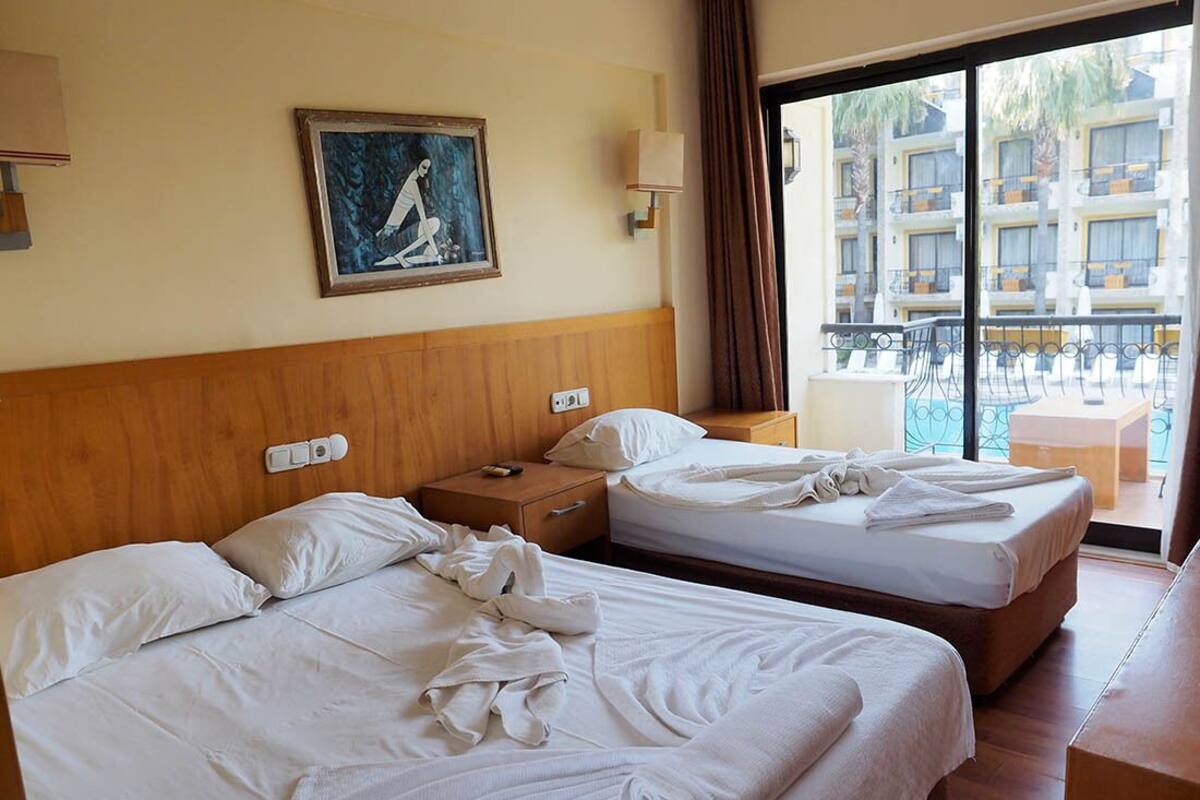 A hotel room with two beds, a painting on the wall, and a balcony with a view of other buildings.