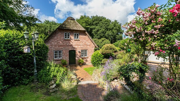 A brick house with a thatched roof, surrounded by lush greenery and flowering plants. - Apartment 'My Little Rose Cottage' with Private Terrace, Private Garden and Wi-Fi (Dörpling)