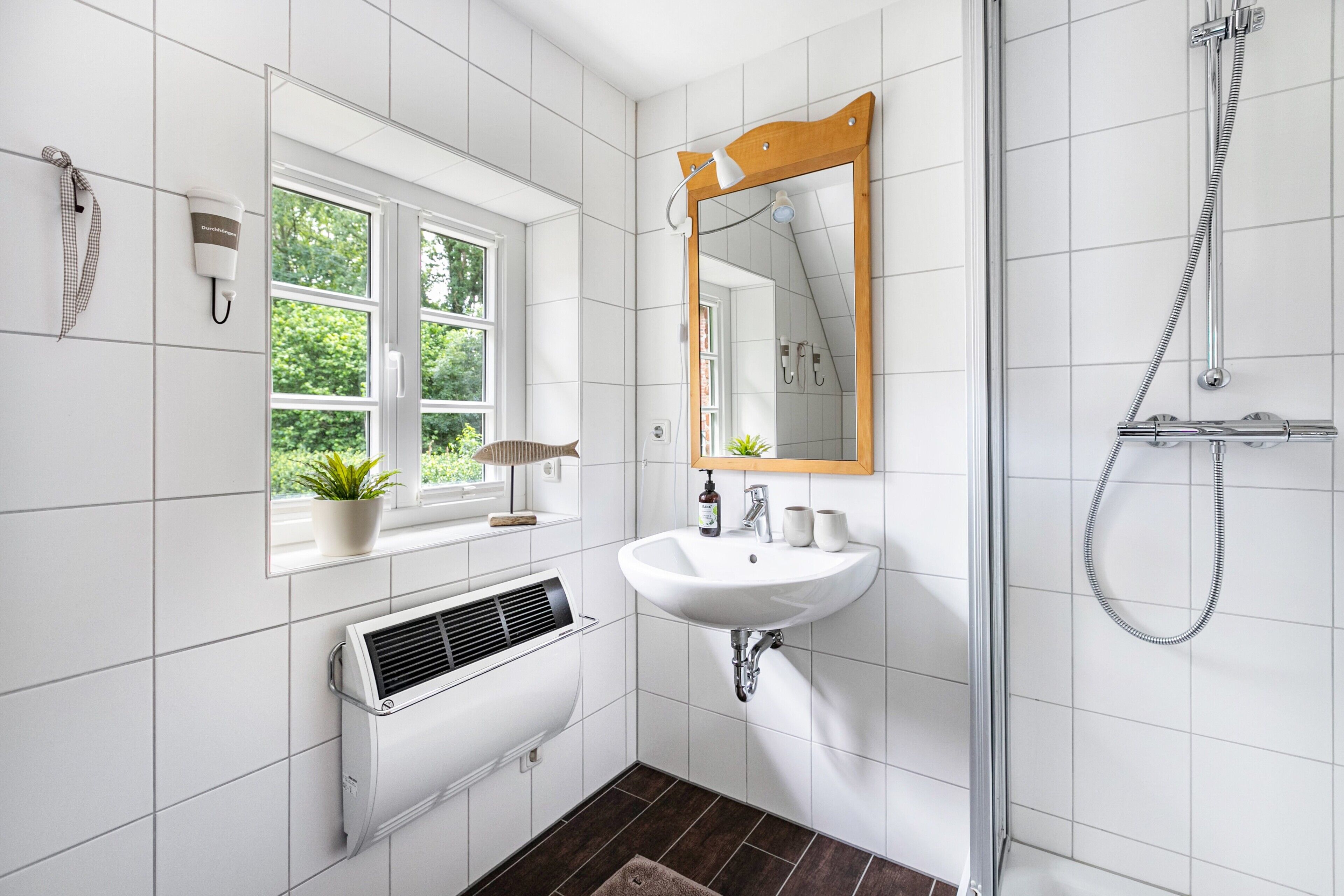 Un baño con azulejos blancos, una ventana con una planta, un lavamanos, un espejo, una ducha y un perchero para toallas.